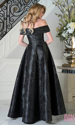A woman in the Marsoni MV1395 off-the-shoulder jacquard long formal dress stands by a staircase and floral arrangement, facing away from the camera.