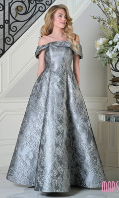 A woman wearing a charcoal off-the-shoulder Marsoni MV1395 long formal dress with a textured floral pattern stands indoors near a staircase and floral arrangement.