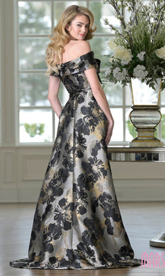 A woman stands indoors by a table with a large floral arrangement, wearing the Marsoni MV1396—a long formal off-the-shoulder gown in black and gold jacquard, perfect for a mother-of-the-bride look.