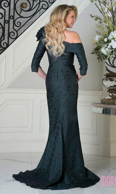 A woman with long wavy blonde hair wears the Marsoni MV1399 long formal dress in black jacquard, standing indoors by a staircase and floral arrangement.