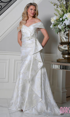 An elegantly dressed woman stands indoors in the ivory Marsoni MV1401 long formal dress with a fitted bodice and draped overlay, near a table topped with a decorative flower arrangement.
