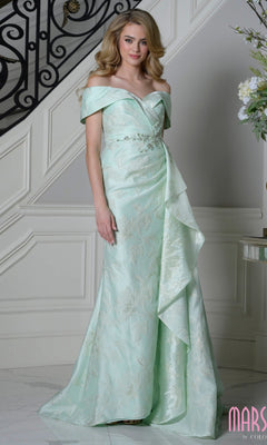 A woman stands indoors near a staircase wearing the Marsoni MV1401 long formal dress in mint. This elegant off-the-shoulder gown features floral patterns, ruffled details, and beautiful textured jacquard fabric for added charm.