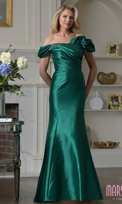 A woman wearing the Marsoni MV1407 long formal dress in emerald, featuring a ruffled off-shoulder design with a ruched bodice and floral accent, stands indoors near shelves and a fireplace.