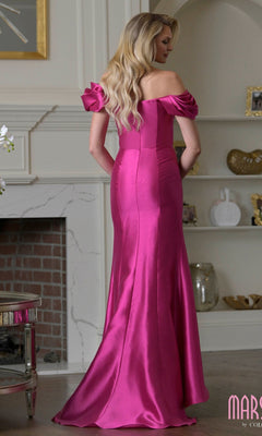 A woman with long blonde hair wears the Marsoni MV1407, a bright pink ruffled off-shoulder satin mermaid gown, standing in a room with a fireplace and shelves.