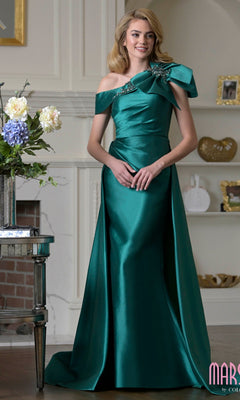 A woman indoors wears the Long Formal Dress: Marsoni MV1408 in emerald, featuring a floor-length taffeta design with an off-the-shoulder asymmetrical neckline and floral shoulder embellishment.