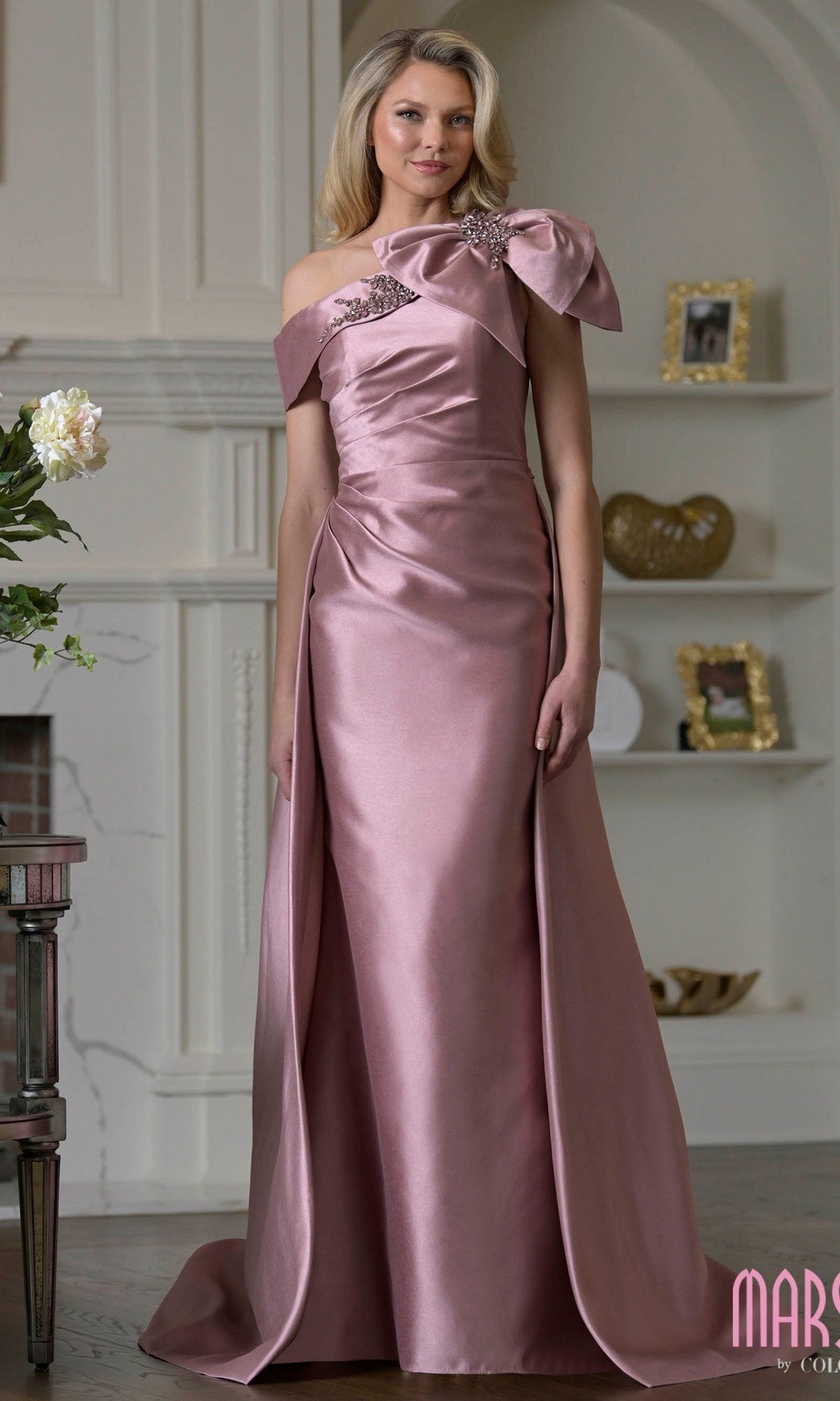 A woman stands in a room wearing the Marsoni MV1408 long formal dress in Rose Wood, featuring taffeta fabric, an asymmetrical neckline, a bold bow accent, and floral embellishment on one shoulder.