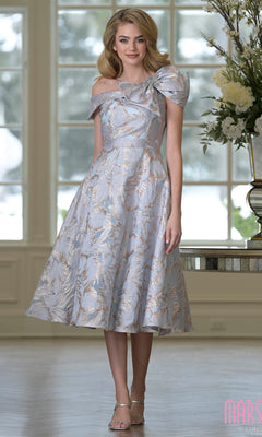 A woman stands indoors by a large window and floral arrangement, wearing the Marsoni MV1413 Knee Length Semi-Formal Dress in Gold Blue with off-the-shoulder style, textured jacquard fabric, and bow detail.