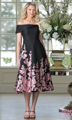 A woman stands indoors in a Marsoni MV1414 knee length semi-formal dress in rose black, paired with black heels and styled with long wavy hair, next to a large floral arrangement.