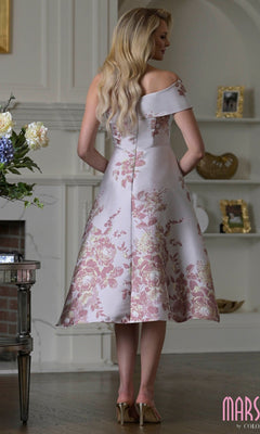 A woman stands indoors with her back to the camera, wearing the Marsoni MV1415 knee-length semi-formal dress, a floral off-the-shoulder fit-and-flare style paired with heels.
