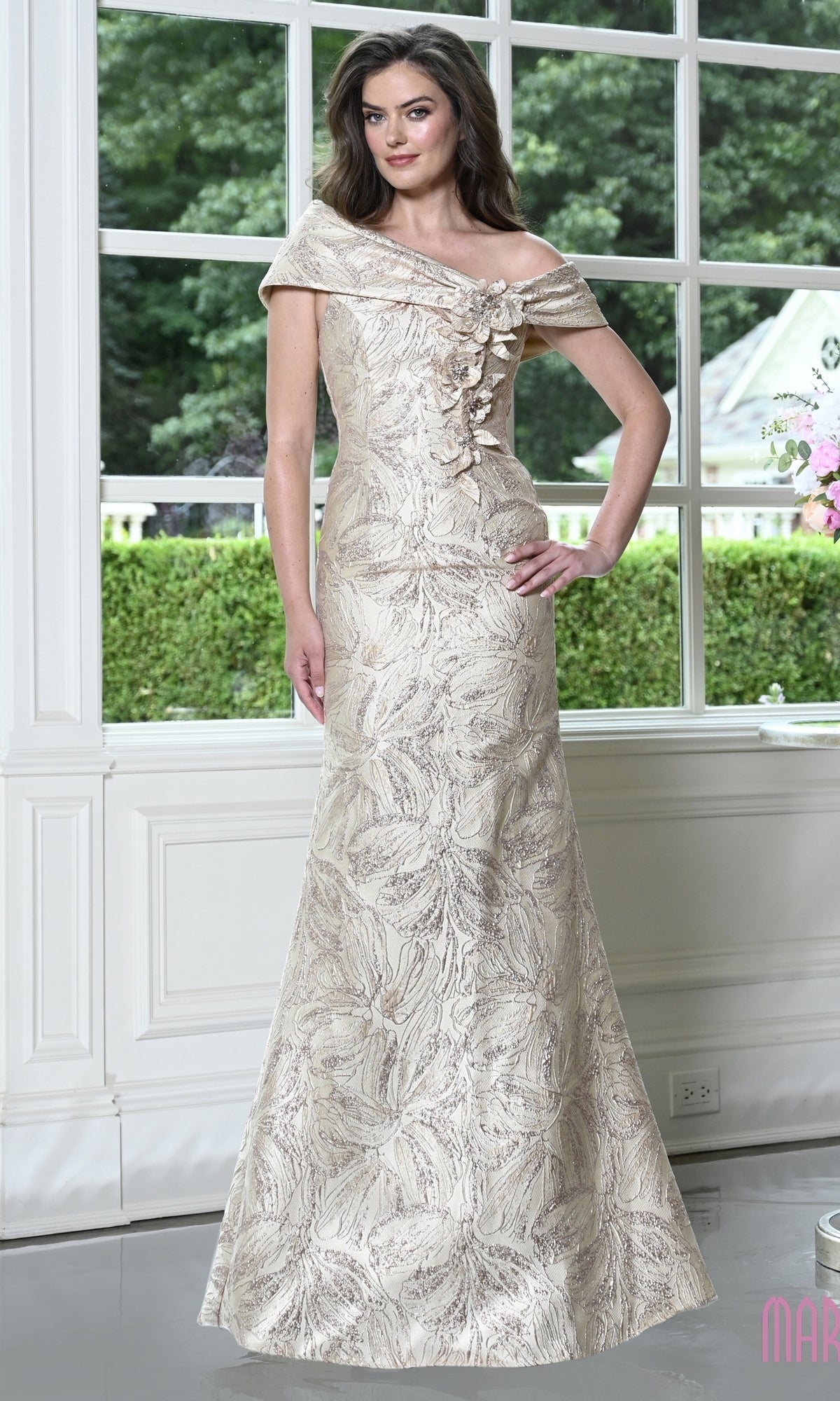 A champagne-colored Marsoni MV1422 floor-length evening gown featuring an off-the-shoulder neckline, bodice embellishments, and an overall metallic floral leaf pattern.