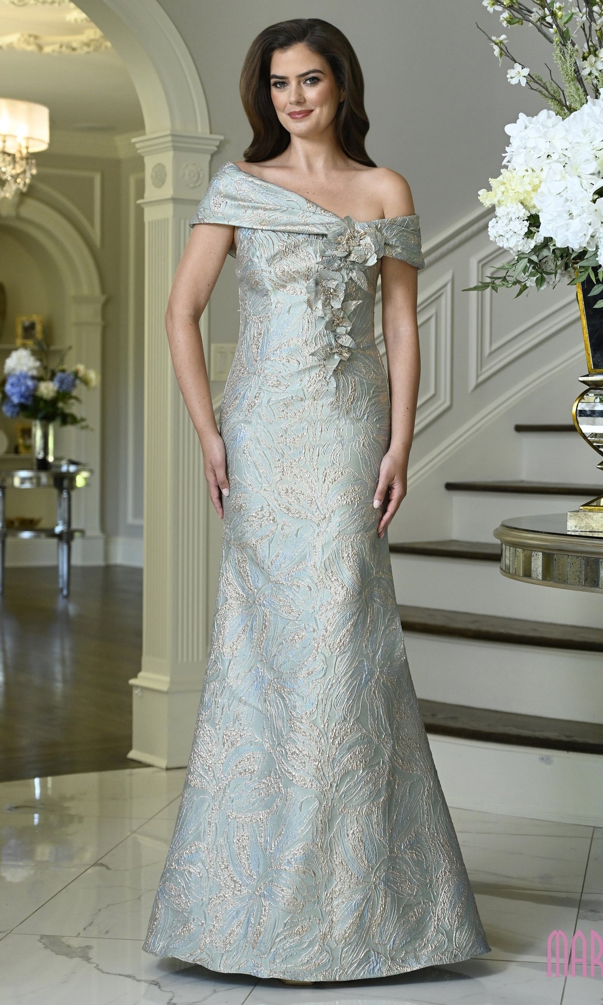 A woman wearing a sage-colored, floor-length Marsoni MV1422 floral evening gown with an off-the-shoulder neckline and ruffled bodice detail.