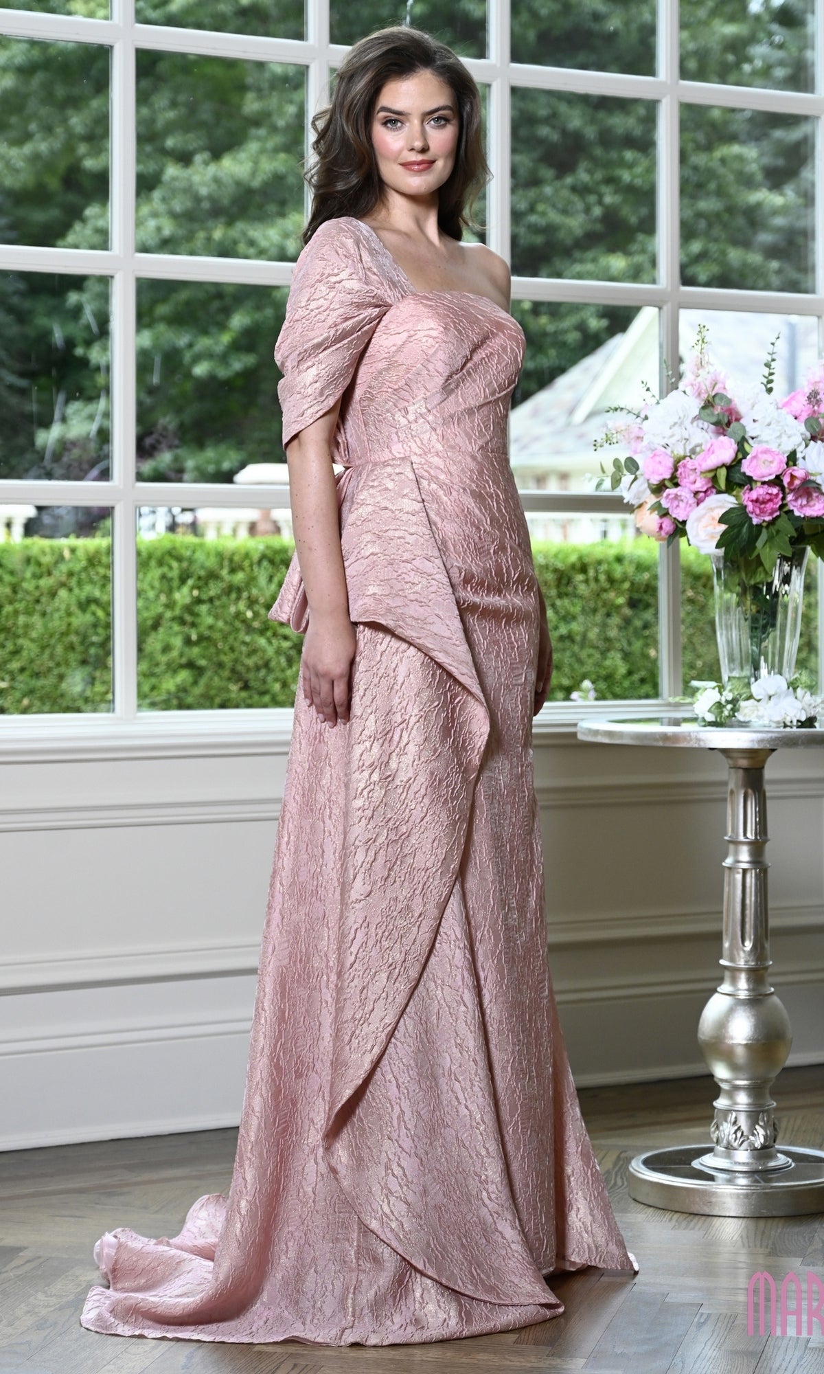 A woman stands wearing a rose-colored, one-shoulder Marsoni MV1437 evening gown with a textured finish and draped waist detail.