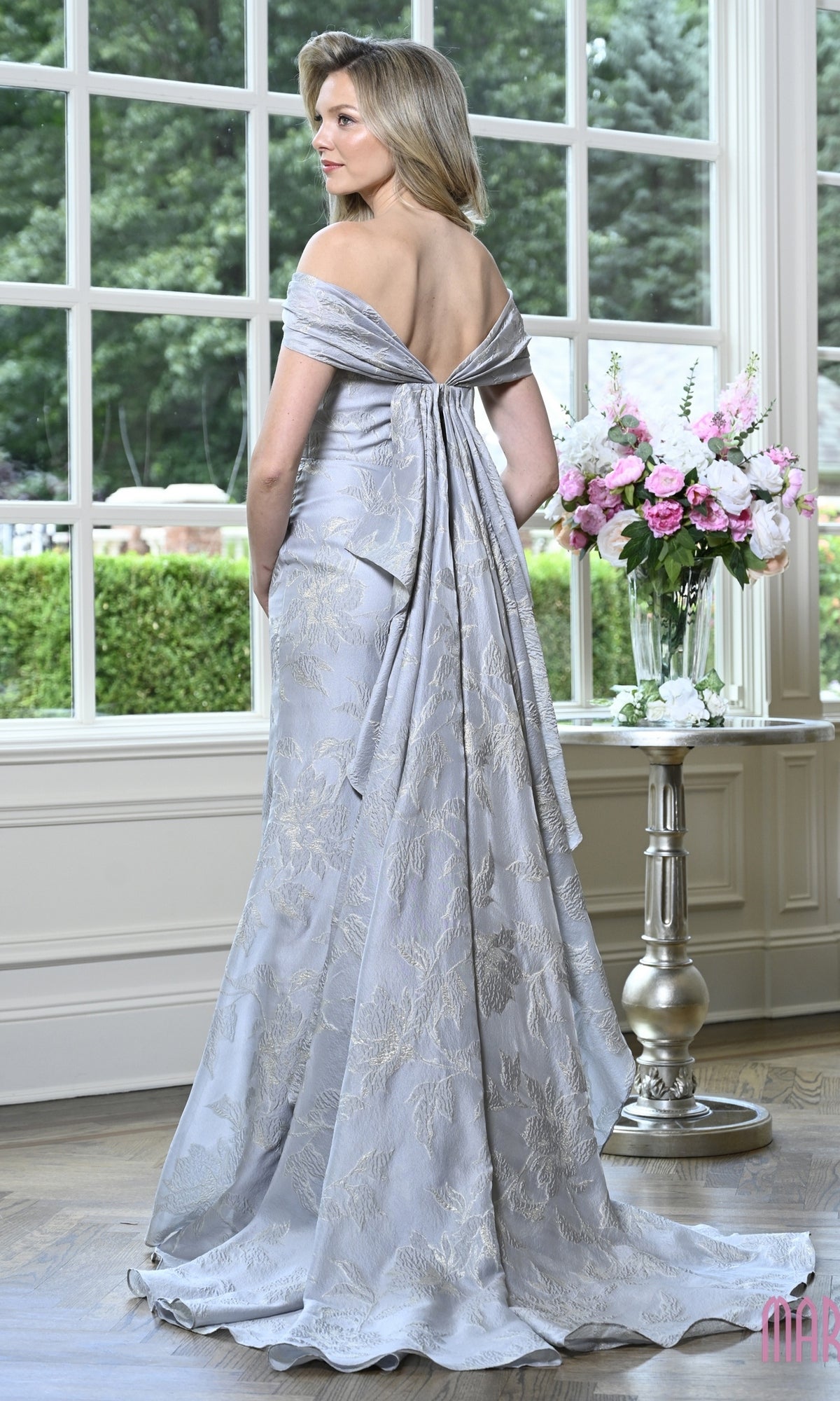 Woman wearing the Long Formal Dress: Marsoni MV1441, a light blue, off-the-shoulder organza jacquard evening gown with a floral pattern and a floor-length, draped back detail.