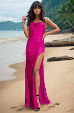 A woman in the Cowl-Neck Long Formal Corset Prom Dress 8035J, featuring a bright pink hue and high slit, stands on a sandy beach with greenery and the ocean in the background.