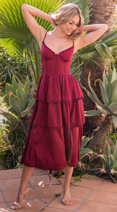 A woman wearing the Velvi Lace-Up Layered Midi Party Dress Maeve stands outdoors on a tiled surface among green plants, lifting her hands to her hair.