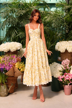 A woman stands indoors among colorful flowers and plants, wearing the Buttercream Floral Casual Midi Dress: Marigold, featuring a buttercream floral print and a corset bodice.