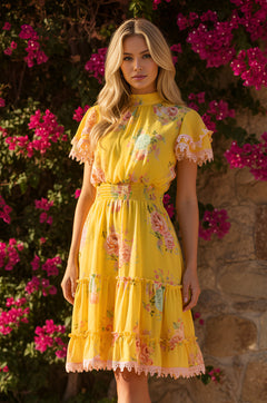 A woman stands by a stone wall with blooming pink flowers, wearing the Sunshine Yellow Short Floral Casual Dress: Maya, featuring lace trim and ruffled sleeves.