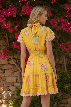 A woman stands outdoors by a stone wall and pink flowers, seen from the back wearing the Maya Sunshine Yellow Short Floral Casual Dress.