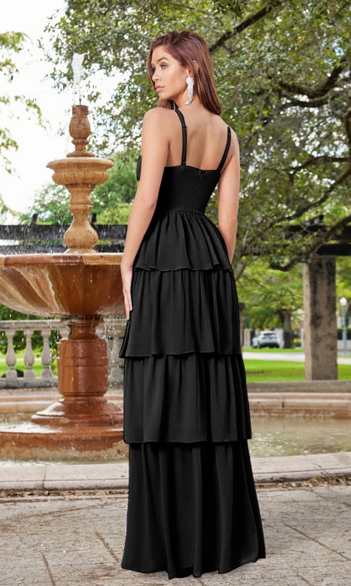 A woman wearing the Velvi Ruffled Long V-Neck Maxi Party Dress Miriam stands outdoors in front of a stone fountain, surrounded by trees and greenery.