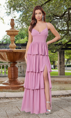 A woman stands by a fountain wearing the Velvi Ruffled Long V-Neck Maxi Party Dress Miriam in lavender, featuring a sleeveless design, high slit, and white heels. Trees and greenery form the background.