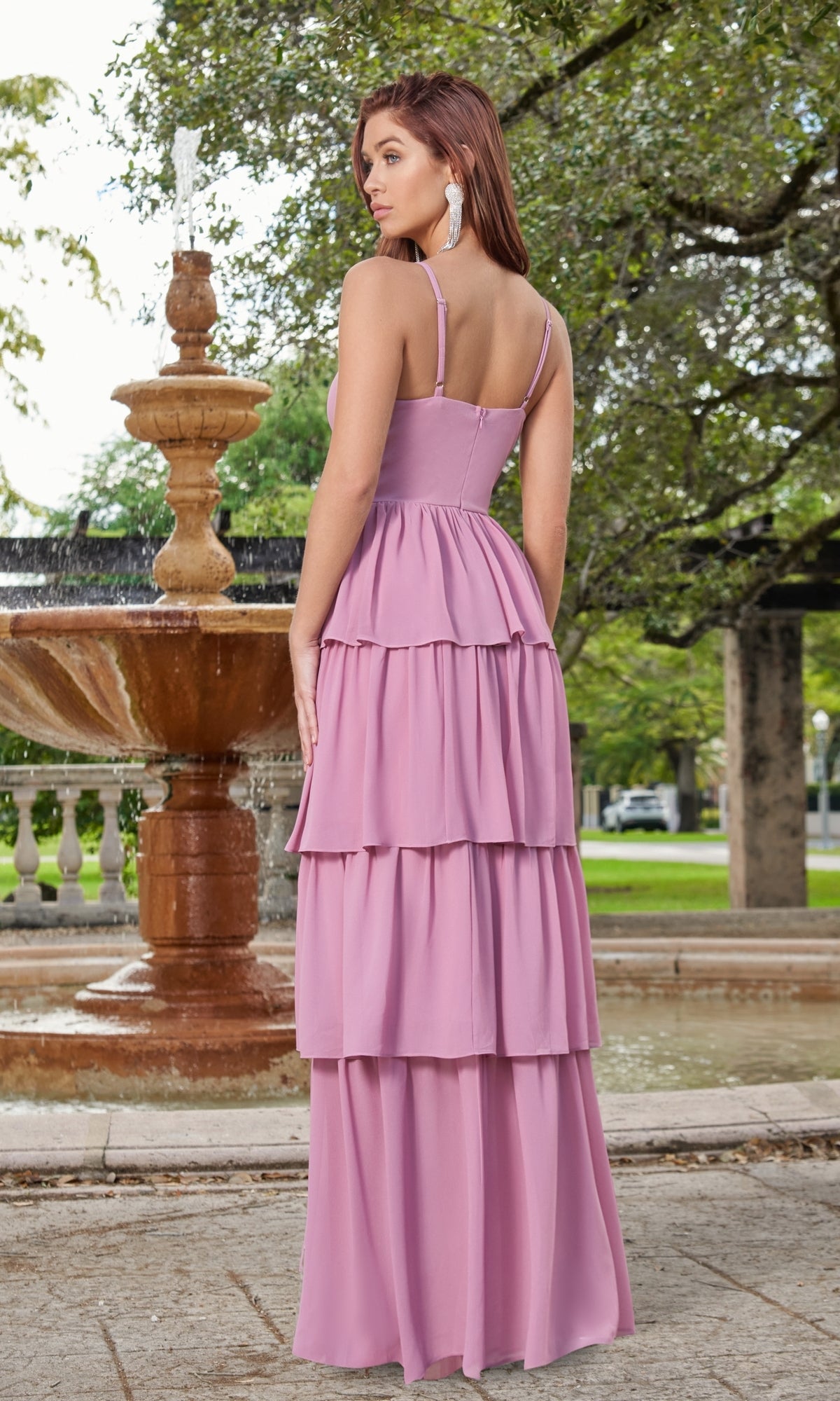 A woman wearing the Velvi Ruffled Long V-Neck Maxi Party Dress Miriam stands outdoors in front of a stone fountain and green trees.