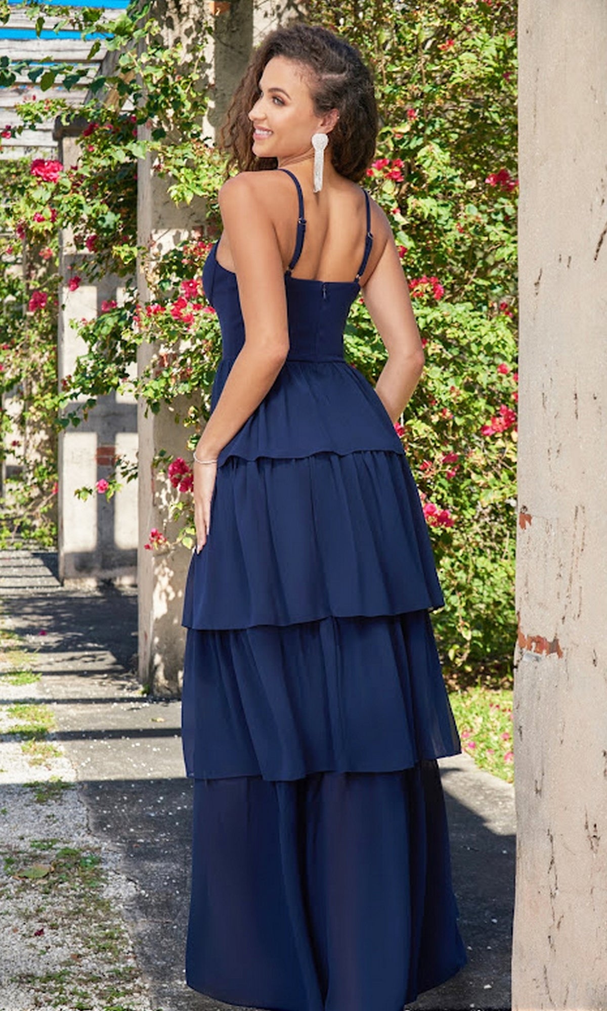 A woman wearing the Velvi Ruffled Long V-Neck Maxi Party Dress Miriam in navy stands outdoors by a stone column and flowering plants, facing away but glancing back over her shoulder.