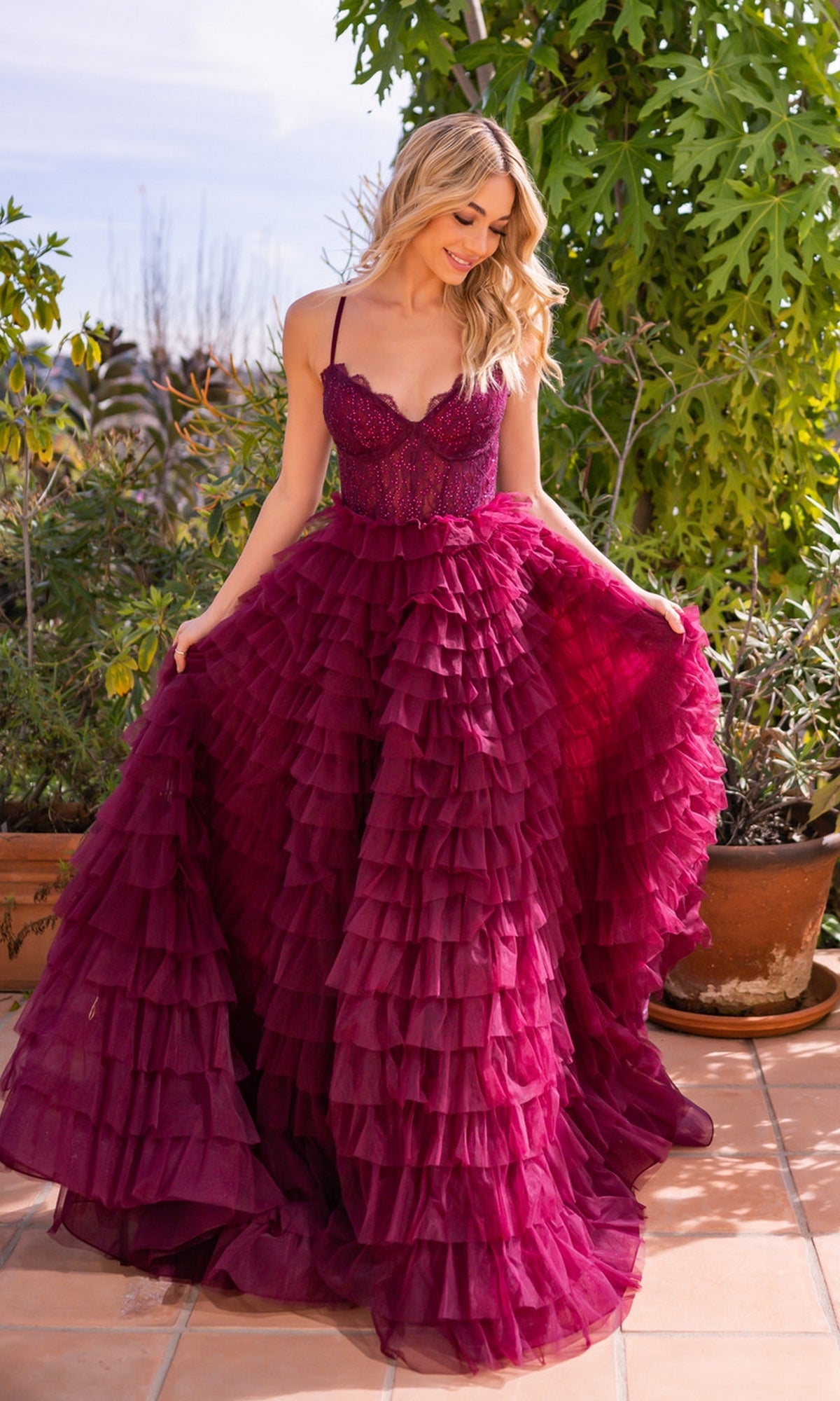 A woman wearing the Velvi Lace-Corset Long Prom Ball Gown Morgan, a deep magenta ruffled dress with spaghetti straps, stands on a tiled patio holding out her gown amid lush potted plants and greenery.