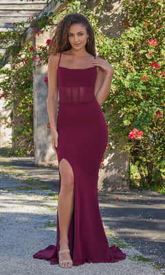 A woman wearing the Velvi Sheer-Waist Long Black Prom Dress Naomi, featuring a sleeveless design, thigh-high slit, and sheer corset waist, stands outdoors near a vine-covered wall.