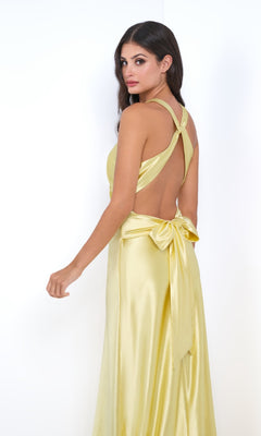 A woman models the Odette by Velvi—a sleeveless, yellow satin long prom dress with a backless design, crisscross straps, empire waist, and large waist bow—against a plain white background.