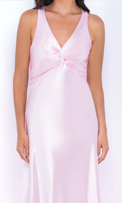 A woman models the Odette by Velvi, a sleeveless satin long prom dress in light pink with a V-neckline, empire waist, and knotted bust detail, standing against a plain white background.