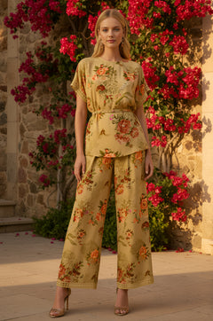 A woman stands outdoors in the Mustard Yellow Floral-Printed Matching Set: Charlotte, featuring wide-leg pants, surrounded by pink flowers and stone walls, embracing vintage-inspired florals.