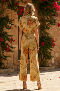 A woman with blonde hair stands outdoors facing away, wearing the Charlotte Mustard Yellow Floral-Printed Matching Set, paired with sandals, with stone walls and pink flowers in the background.