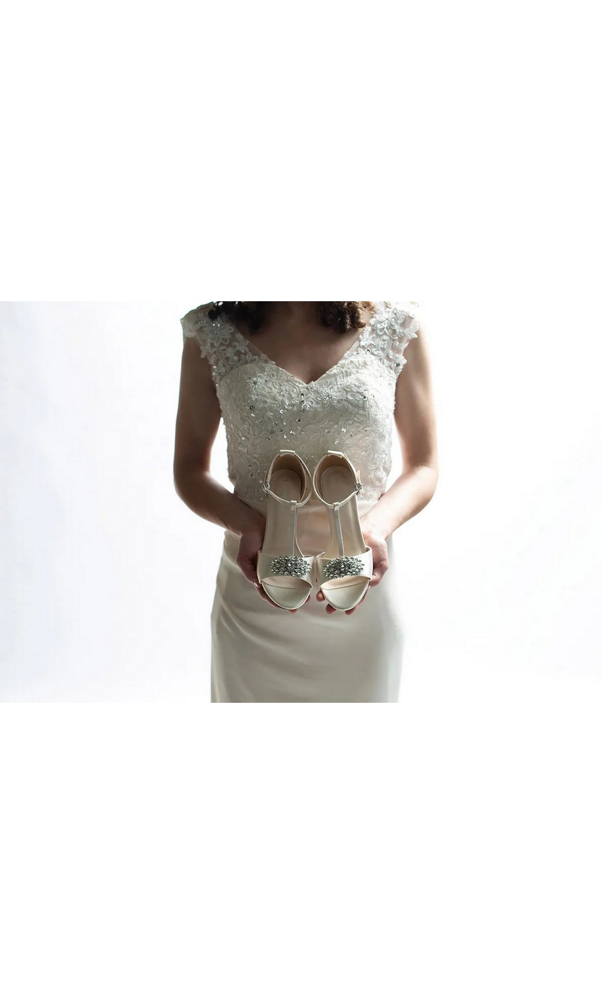 A woman in a white wedding dress holds up Ophelia White Prom Shoes: Dyeables 4468, a pair of white satin kitten heel sandals featuring jeweled embellishments on the toes.
