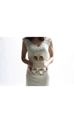 A woman in a white wedding dress holds up Ophelia White Prom Shoes: Dyeables 4468, a pair of white satin kitten heel sandals featuring jeweled embellishments on the toes.