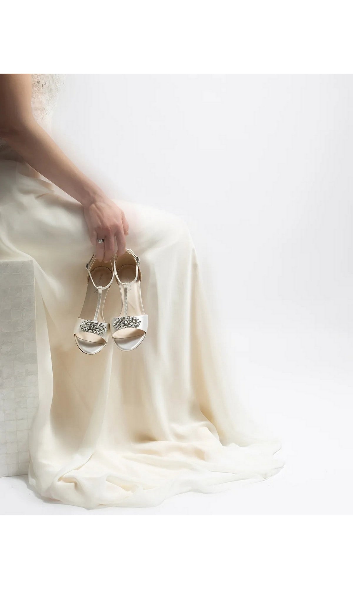 A person in a long white gown holds a pair of white satin Ophelia White Prom Shoes: Dyeables 4468 with rhinestone-embellished T-straps.