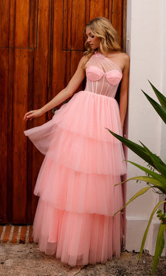 A woman in the Sheer-Bodice Long Layered Prom Ball Gown P1400, featuring a light pink tiered skirt and fitted corset-style bodice, stands by a wooden door and looks down at her dress.