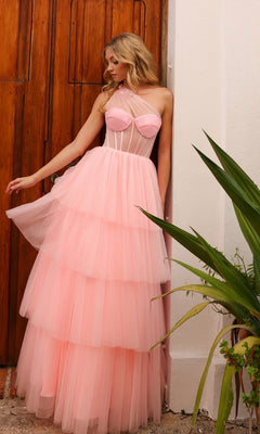 A woman wearing the Sheer-Bodice Long Layered Prom Ball Gown P1400—a light pink, strapless, tiered tulle dress—stands by a wooden door, gazing to the side with green plants in the foreground.