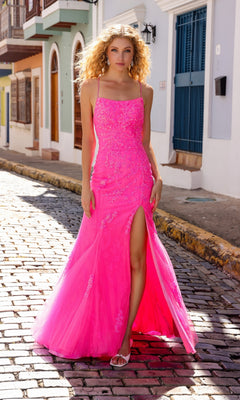 A woman in the Sequin-Floral Long Lace Prom Dress P1401, a bright pink gown with a thigh-high slit and sequin floral designs, stands on a cobblestone street lined with colorful buildings.