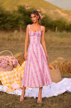 A woman wears the Lilac Floral Casual Midi Dress: Celenia, standing on a picnic setup with a basket, flowers, and checkered blanket—an ideal summer wedding guest dress for a grassy outdoor event.