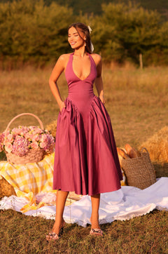 Wearing the Solena Vintage Rose Purple Midi Casual Dress, a woman stands outdoors on a picnic blanket beside a basket of flowers and bread, with hay bales in the background.