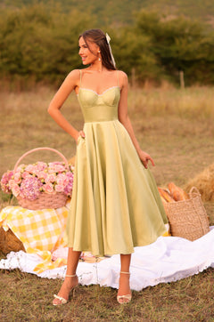 A woman in the Olivetta Olive Green Satin Midi Wedding Guest Dress stands outdoors on a picnic setup with flowers, bread, and a yellow-checkered cloth on the grass.