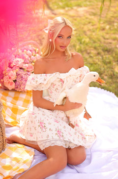 A woman in the Marella Cream White Floral Short Graduation Dress sits on a picnic blanket outdoors, holding a white duck, with a basket and pink flowers nearby.