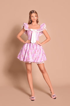 A woman poses against a beige backdrop in the Marella Lilac Floral Off-Shoulder Mini Casual Dress with ruffled details and a white bow, styled perfectly with pink bow-adorned heels.