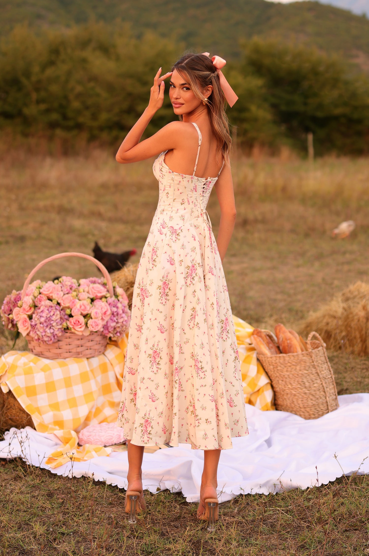 A woman wearing the Cream White Floral Casual Midi Dress: Rosella stands outdoors on a picnic blanket, with a basket of flowers and bread beside her and green hills in the background.