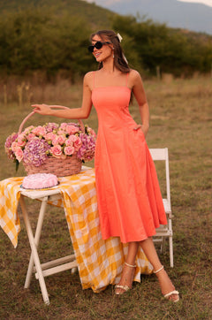 A woman wearing the Papaya Midi Casual Summer Dress in Solid Papaya Punch stands outdoors by a table with a basket of pink flowers, a yellow checkered cloth, and a folded scarf. She smiles and wears sunglasses.