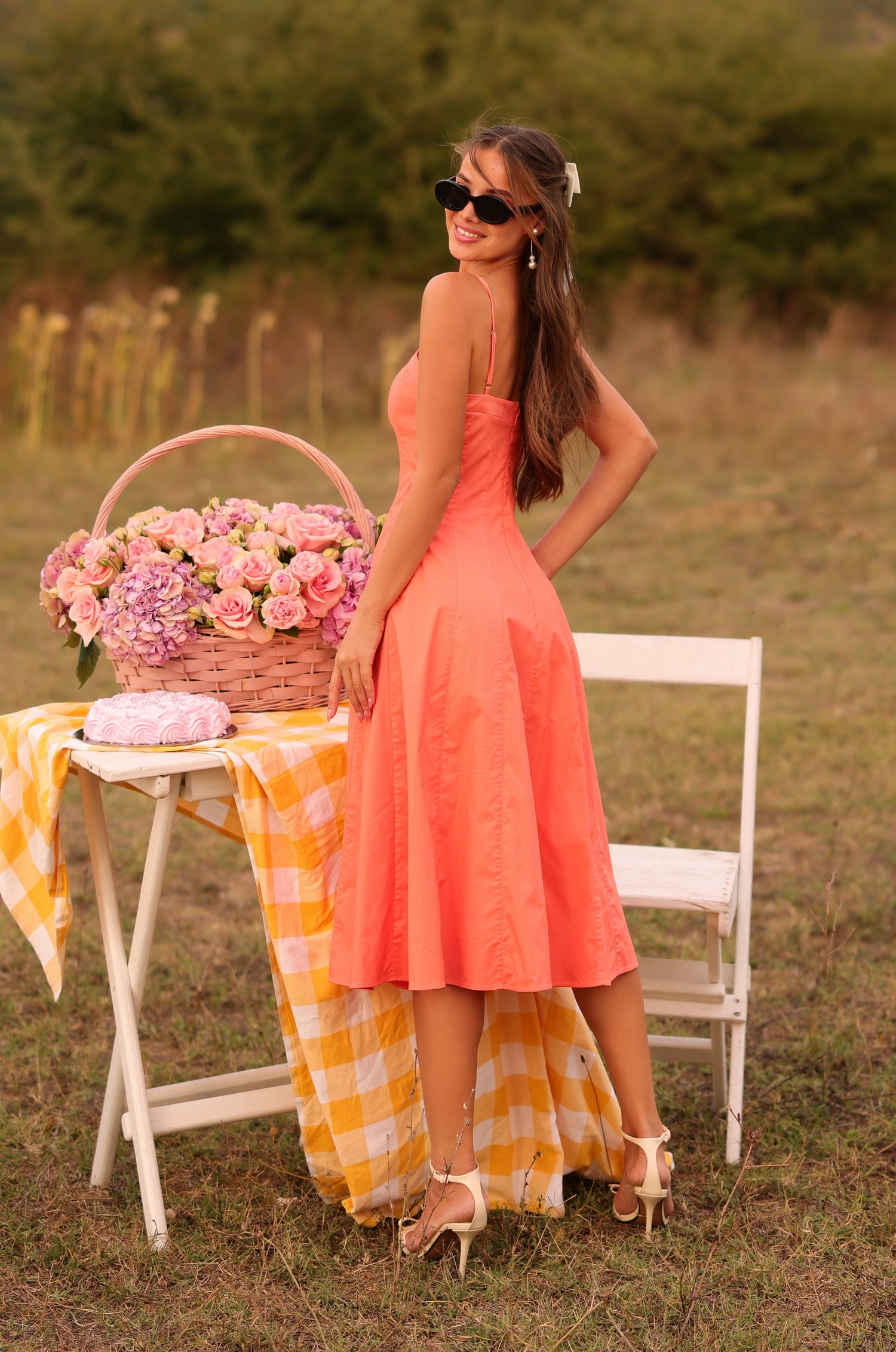A woman in the Papaya Midi Casual Summer Dress: Auberine and heels stands by a table with a yellow checkered cloth, a basket of flowers, and a folded blanket outdoors.