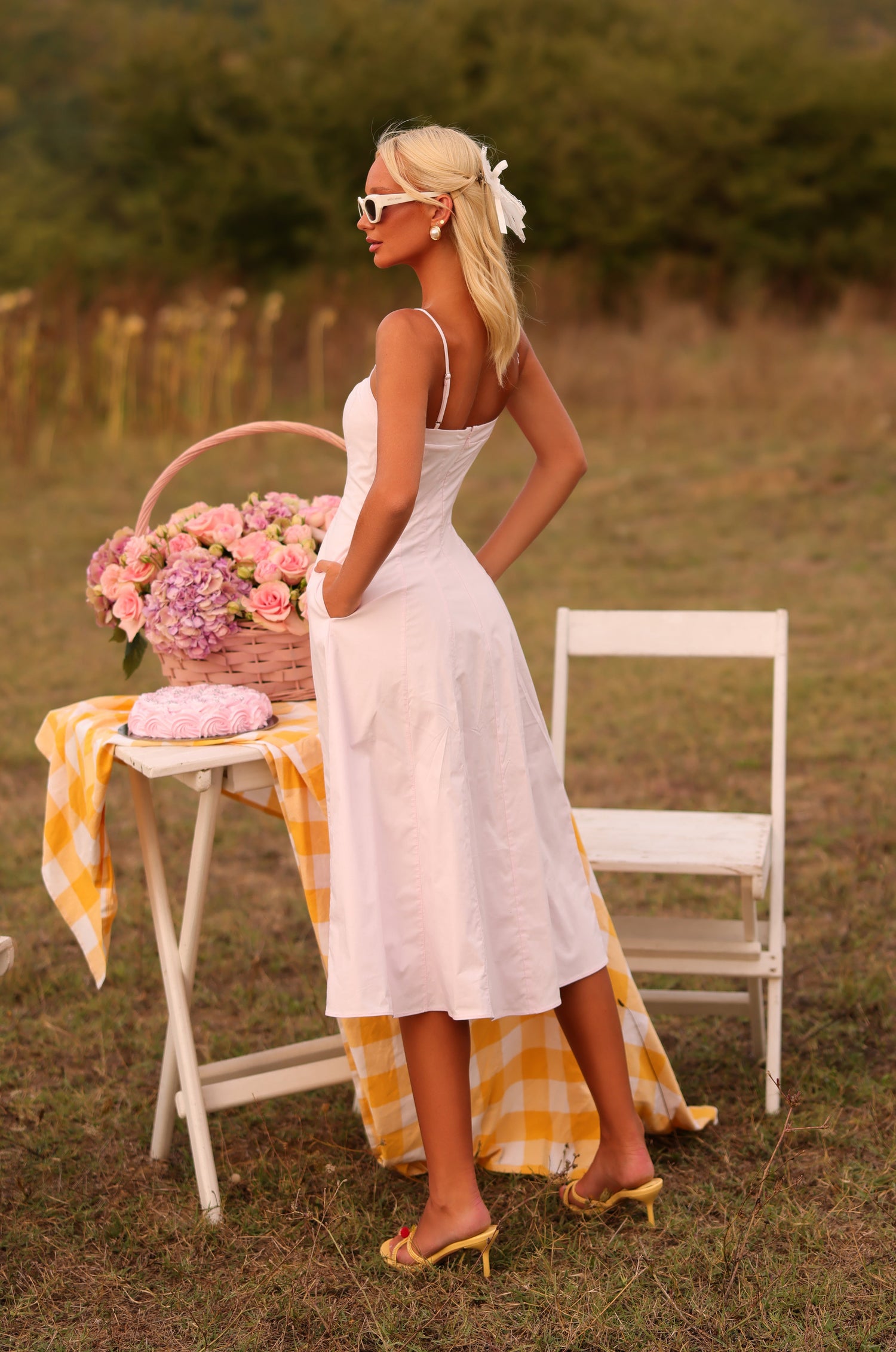 A woman in a Primrose Pink Simple Midi Casual Dress: Auberine with hidden side pockets stands outdoors by a table with flowers, a basket, and cake, surrounded by white chairs and a yellow checked cloth.