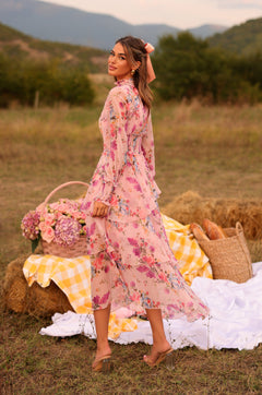 Wearing the Bellaria Long Sleeve Blush Pink Floral Maxi Dress, a woman stands on grass by a picnic setup with hay bales, flowers, and bread, surrounded by trees and mountains.