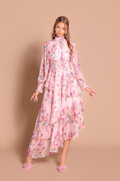 A woman wears the Long Sleeve Blush Pink Floral Maxi Dress: Bellaria, featuring layered ruffles and an asymmetrical hem—perfect for wedding guests—standing against a beige background.