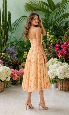 A woman in the Yellow Floral Midi Casual Dress: Millie and nude heels stands indoors, surrounded by colorful flowers and green plants in woven baskets.